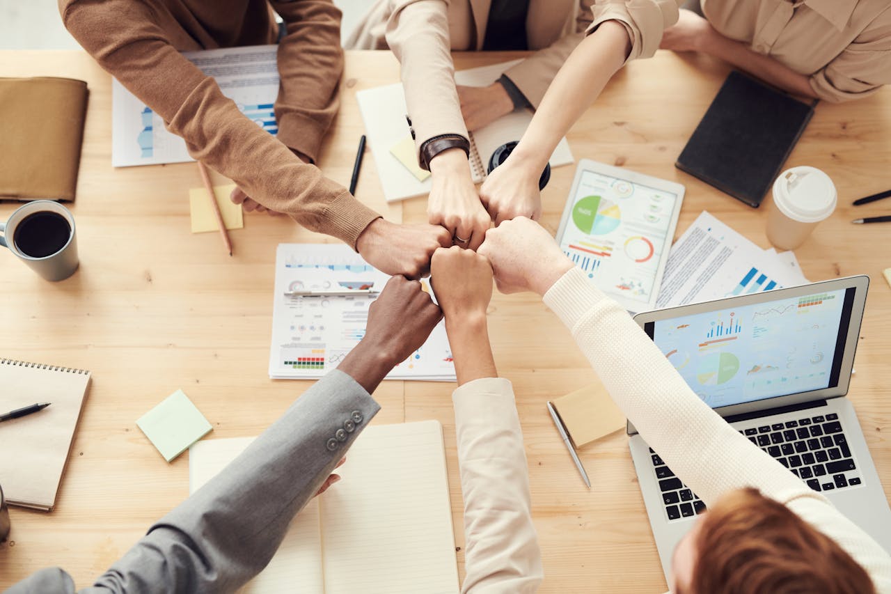 Top view of a diverse team fist bumping over a meeting table with paperwork and laptops, symbolizing teamwork.